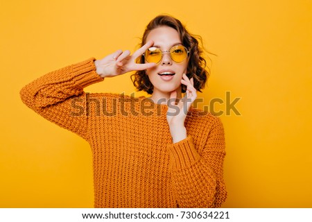 Close-up portrait of charming young woman with gently smile posing with peace sign in trendy glasses. Stylish girl in bright sweater having fun during photoshoot in studio.