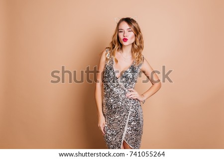 Elegant caucasian woman with curly hairstyle posing with kissing face expression at  party. Charming birthday girl in sparkle dress standing in confident pose on light background.