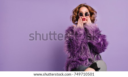 Spectacular young woman with trendy makeup posing with surprised face expression on bright purple background. Indoor photo of shocked female model in stylish fur jacket.
