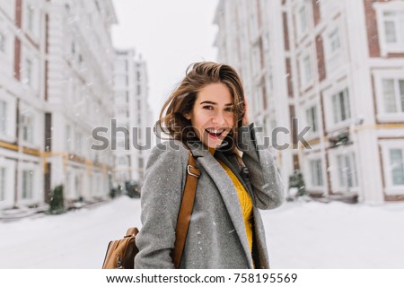 Close-up portrait of ecstatic woman in elegant gray coat standing on the street in snowy day. Outdoor photo of fashionable female model with brown bag walking around city in winter vacation.