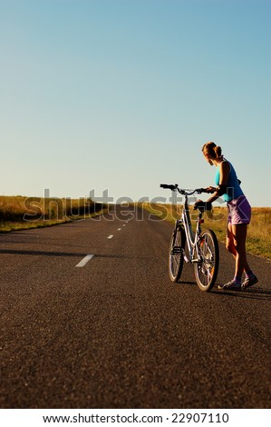 Girl with bicycle on the non-urban road in the sunset.