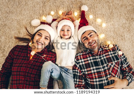 Christmas. Family. Happiness. Top view of dad, mom and daughter in Santa hats looking at camera and smiling while lying on the floor at home