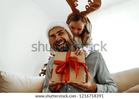 Christmas. Family. Love. Little girl is covering her dad\'s eyes making a surprise with a gift box. Near the Christmas tree at home