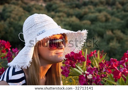 Young smiling blond caucasian white woman with hat and sunglasses smelling red flowers.