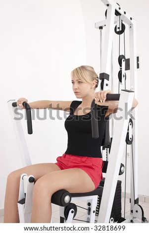 Young blond caucasian woman working out in gym. Pumping iron while sitting, lifting weight, pushing exercise. 3/4 view. Serious facial expression. Model looks aside. Closeup.
