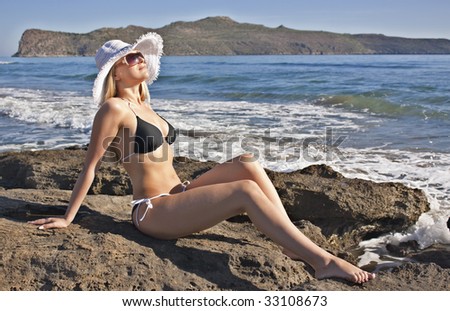 Young smiling blond caucasian white woman wearing black bikini, hat and sunglasses.