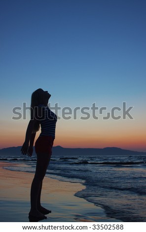 Silhouette of a young woman standing on a beach at sunset. Model looks up. Side view.