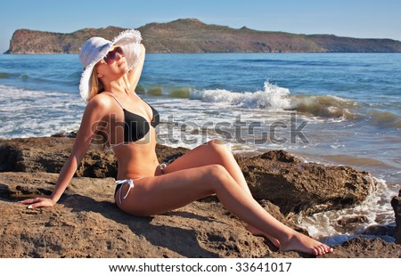 Young smiling blond caucasian white woman wearing black bikini, hat and sunglasses. Model lying on a beach, looking aside to the sun.