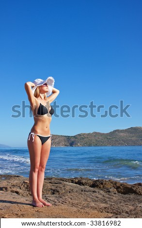 Young smiling blond caucasian white woman wearing black bikini, hat and sunglasses. Model standing, looking aside to the sun.