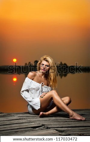 beautiful girl with a white shirt on the pier at sunset.Sexy  woman with long legs  sitting on a pier .Color image of a beauty girl sitting on a pier, overlooking a lake