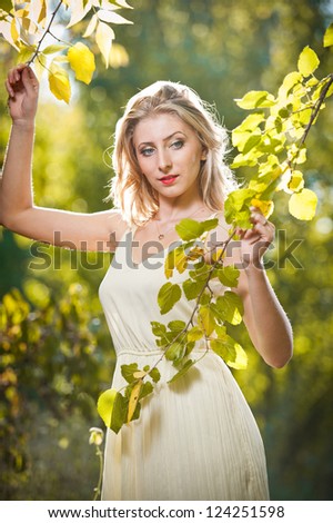 Young attractive woman in a romantic autumn scenery. Blonde woman posing in white dress in autumn scenery.Girl with leaves in hand and fall yellow maple garden background