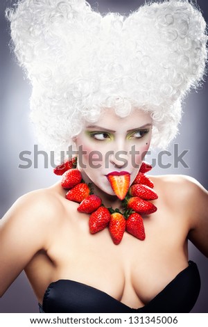 Creative makeup beauty shot of model with strawberries, artistic edit .Woman with strawberry necklace, wig and makeup professionally posing in studio.Beauty with  strawberry in her mouth