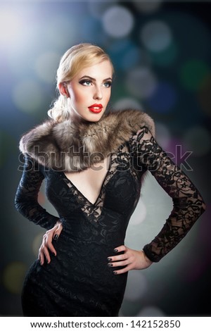 Attractive  blonde woman in black lace dress and fur posing in studio. Photo of fashionable elegant brunette woman posing, looking forward.sensual woman in dress with cleavage.Woman in elegant  dress