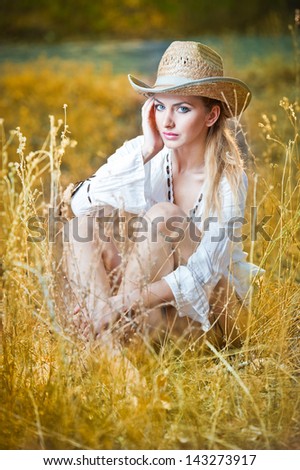 fashion portrait woman with hat and white shirt sitting on a hay stack.very cute blond woman sitting down outdoor on the yellow grass with a hat