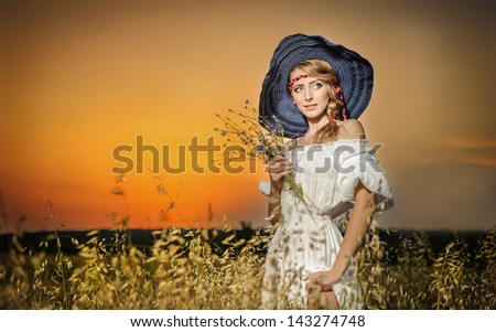 Young woman standing on a wheat field with sunrise on the background.Portrait of girl in field.Romantic young woman posing outdoor.Attractive woman in white dress in yellow wheat field at sunrise.