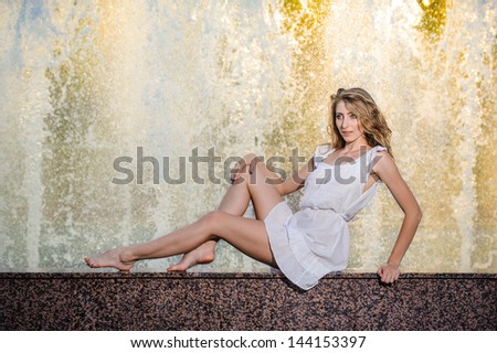 Attractive girl in white short dress sitting on parapet near the fountain in the summer hottest day.Girl with dress partly wet playing with water.Portrait of beautiful blonde women near the fountain