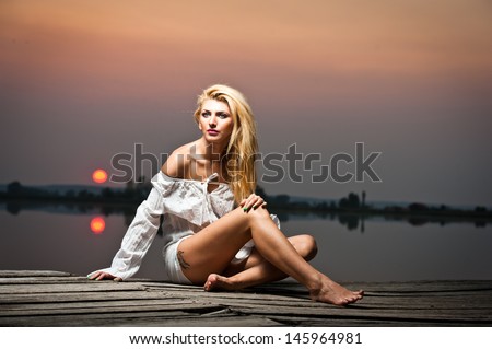 beautiful girl with a white shirt on the pier at sunset.Sexy woman with long legs sitting on a pier .Color image of a beauty girl sitting on a pier, overlooking a lake