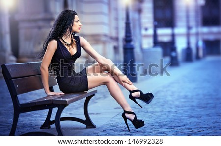 handsome attractive girl wearing short skirt and high heels standing outside in urban scene.Fashion model in blue short skirt with long sexy legs on the street .Woman sitting on bench in the city