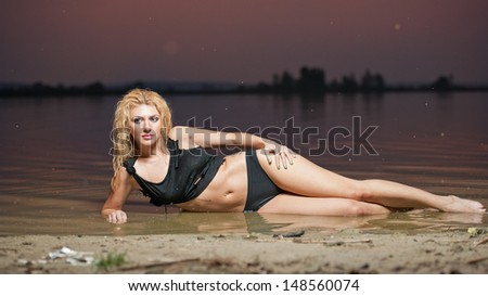Sexy blonde woman in water at sunset .Beautiful swimsuit model.The beautiful sensual bikini model posing against a setting sun on a body of water .Erotic art photo