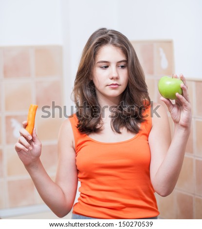 Pretty teen girl looking at a carrot and an green apple, indoor. Beautiful teenage girl holding a green apple and a carrot in her hands.