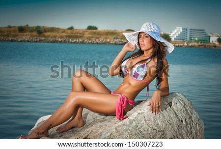 fashion portrait of young sexy brunette girl in bikini and big hat at the beach.Sensual attractive woman in water wearing bikini and nice tits. Woman with perfect body relaxing on the rock.
