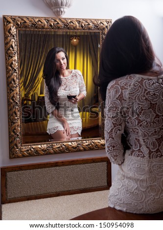 Beautiful girl in a short white dress with glass of wine looking into mirror.Sensual woman wearing a white short dress in the old hotel.Sensual elegant young woman in white dress looking into mirror