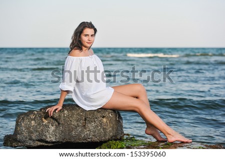 caucasian teen girl in bikini and white shirt  lounging on lava rocks by the ocean .Teenage girl in white shirt sitting on a rock in the sea .Beautiful girl relaxing on the rock near black  sea.