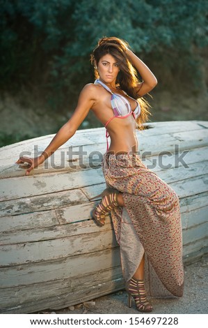Fashion portrait of  sexy brunette  girl in swimsuit leaning on a wooden boat.Sensual attractive woman with long skirt posing outdoor. Woman with perfect body and long hair relaxing on the old boat