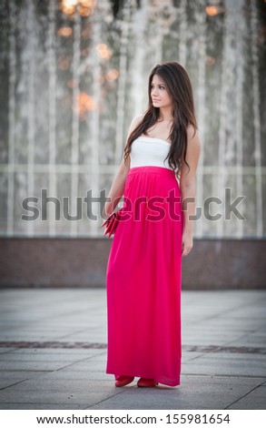 Full length of young caucasian female with long red skirt standing near fountain.Romantic portrait of the woman.