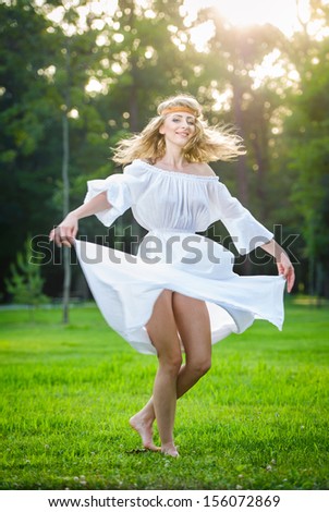 Young woman standing in a park. Portrait of girl in a meadow. Romantic young woman posing outdoor. Attractive woman in long white dress with a flower power look