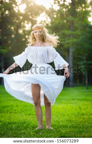 Young woman dancing in a park. Girl in a meadow dancing on green grass. Romantic young woman posing outdoor. Attractive flower power look woman in long white dress
