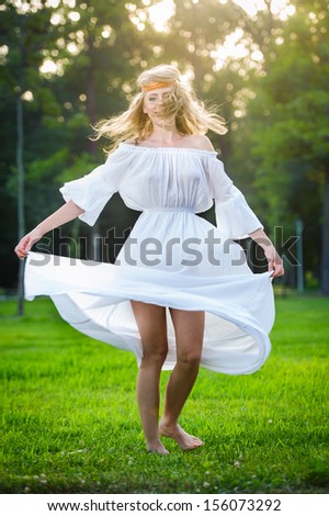 Young woman dancing in a park. Girl in a meadow dancing on green grass. Romantic young woman posing outdoor. Attractive flower power look woman in long white dress