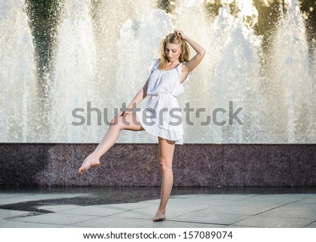 Attractive girl in white short dress sitting in front of a fountain in the summer hottest day. Girl with dress partly wet dancing. Beautiful blonde women near the fountain in a ballet position