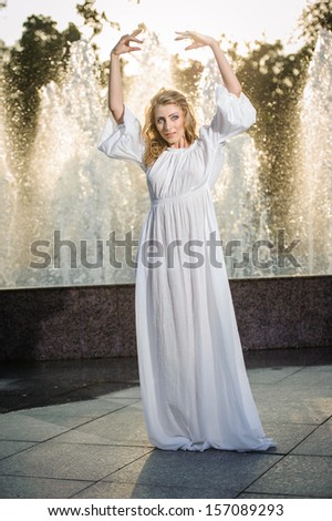 Attractive girl in white long dress sitting in front of a fountain in the summer hottest day. Girl with dress partly wet posing near a fountain. Blonde women near the fountain in a ballet position