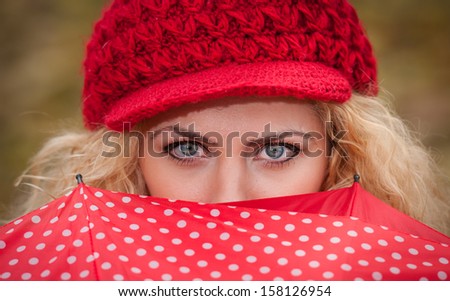 Beautiful blue eyes over colorful umbrella. Attractive blonde girl with red cap looking over red umbrella outdoor shoot. Attractive young woman in a autumn fashion shoot.