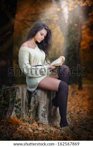 Young Caucasian sensual woman reading a book in a romantic autumn scenery. Portrait of pretty young girl in the forest in autumn day. Fashion portrait of a beautiful young woman in autumn forest