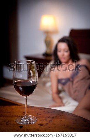Mysterious lady laying in bed with a glass of red wine foreground. Sensual woman on bed and glass of wine. Beautiful girl in sensual lingerie posing provocatively indoor,  relaxation moments