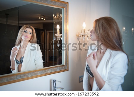 Sensual elegant woman in office outfit looking into a large mirror. Beautiful and sexy blonde young woman wearing an elegant white jacket posing in a mirror. Fashionable model.