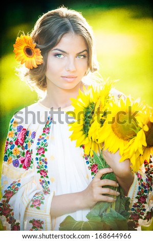 Young girl wearing Romanian traditional blouse holding sunflowers outdoor shot. Portrait of beautiful blonde girl with bright yellow flowers bouquet. Beautiful woman with yellow flower in hair posing