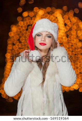 Fashionable lady wearing Xmas hat and white fur coat outdoor. Portrait of young beautiful woman in winter style. Bright picture of beautiful blonde woman with make up wearing Santa hat