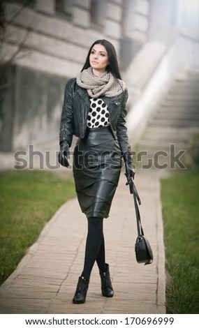 Attractive Young Woman In A Winter Fashion Shot. Beautiful Fashionable Young Girl In Black Leather Waking On Avenue. Elegant Long Hair Brunette With Handbag And Scarf In Urban Scenery.