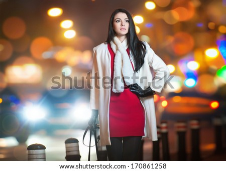 Fashionable lady wearing red dress and white coat outdoor in urban scenery with city lights in background. Full length portrait of young beautiful elegant woman posing in winter style. Street shot.