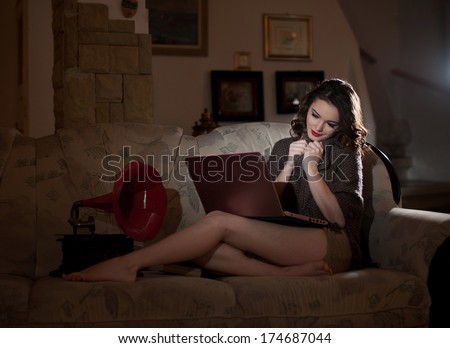 Beautiful young woman sitting on bed working on laptop having a red gramophone near her, in boudoir scenery. Attractive brunette girl with long hair and long legs laying down on bed with a laptop