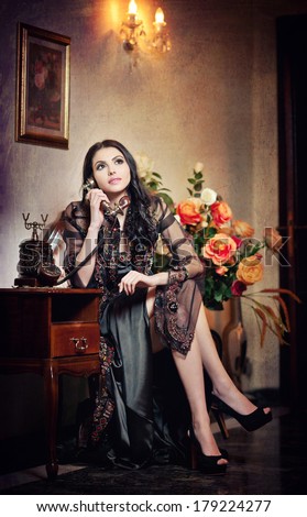 Young beautiful luxurious woman in long elegant dress talking by phone in a elegant scenery. Seductive brunette woman with roses in background holding a vintage phone in luxurious classic interior