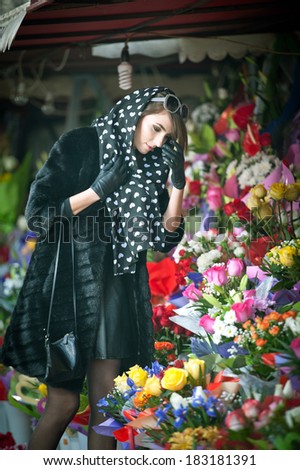Beautiful brunette woman with gloves choosing flowers at the florist shop. Fashionable female with sunglasses and head scarf at flower shop. Pretty brunette in black choosing flowers - urban shot