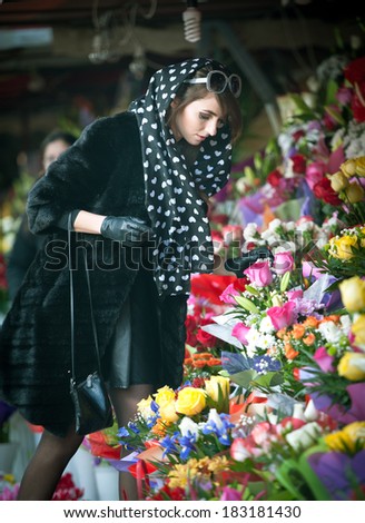Beautiful brunette woman with gloves choosing flowers at the florist shop. Fashionable female with sunglasses and head scarf at flower shop. Pretty brunette in black choosing flowers - urban shot
