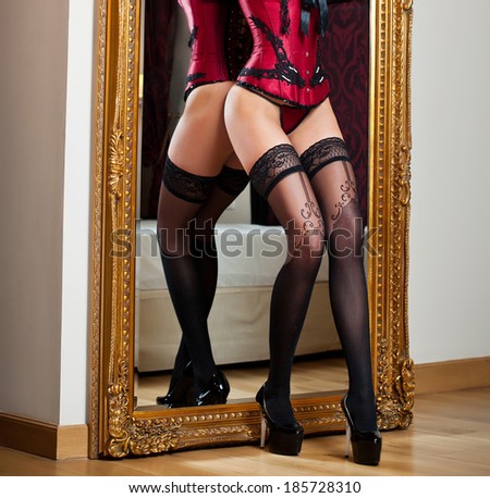 Attractive woman in red lingerie posing challenging near a large mirror. Sensual woman with long legs and high heels in corset. Classic boudoir shot. Erotic photo.