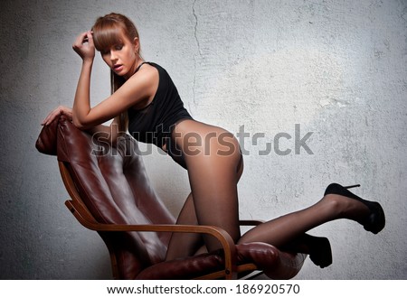 Attractive red hair model with black corset sitting provocatively on chair - gray background. Fashion portrait of a sensual woman - studio shot. Beautiful redhead female in black posing provocatively.