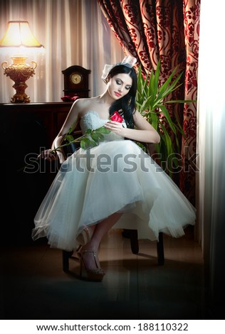 Young beautiful luxurious bride sitting holding a red rose in classic scenery. Attractive bride with curtains and lamp in background. Seductive brunette with ribbon in luxury manor, vintage style