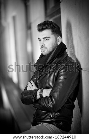 Top view of handsome male model in leather jacket posing thinking. Cool and trendy young man standing against wall. Black and white photo of attractive young man.